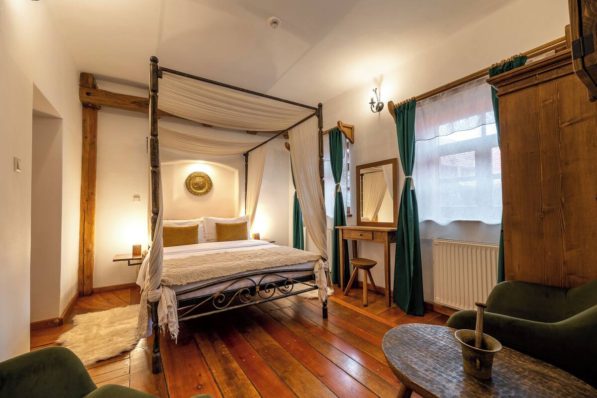 A four-poster bed with a canopy, a wooden desk with a chair, a green armchair, and a small table with a candle.