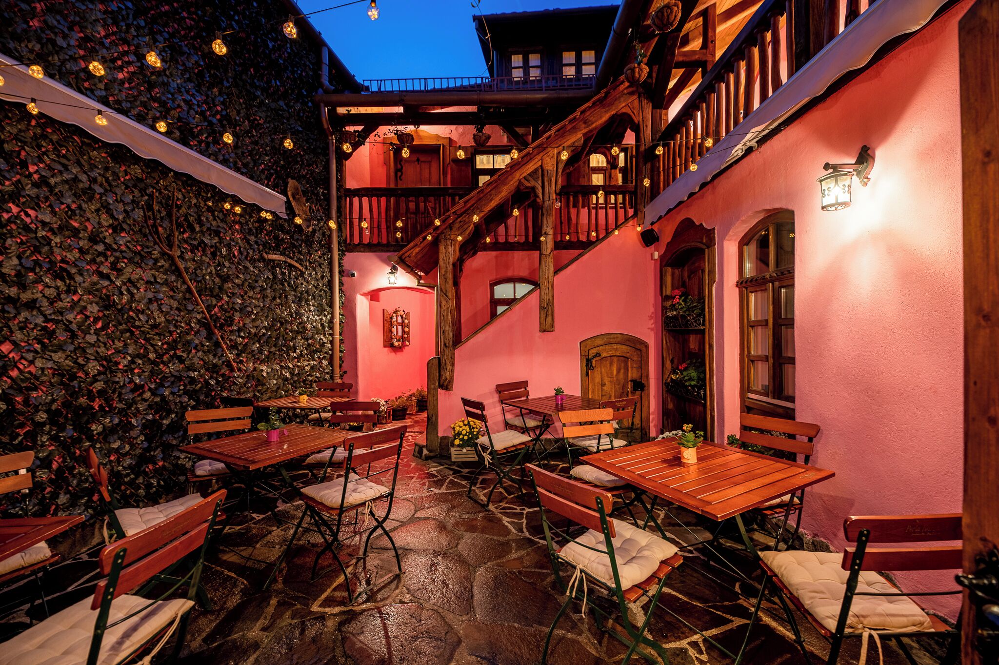 Un patio exterior con mesas y bancos de madera, suelo de piedra y un edificio de color rosa con balcones y escaleras de madera.