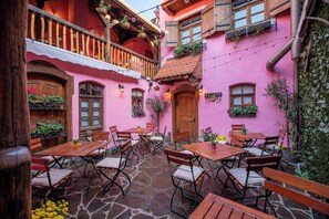 Un patio con mesas y sillas de madera, suelo de piedra y un edificio rosa con balcones y ventanas de madera.