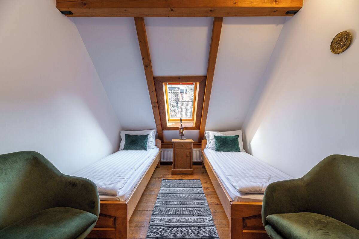 A small, cozy room with two single beds, a wooden chest of drawers, a small table with a lamp, and a window with a view of a building outside.