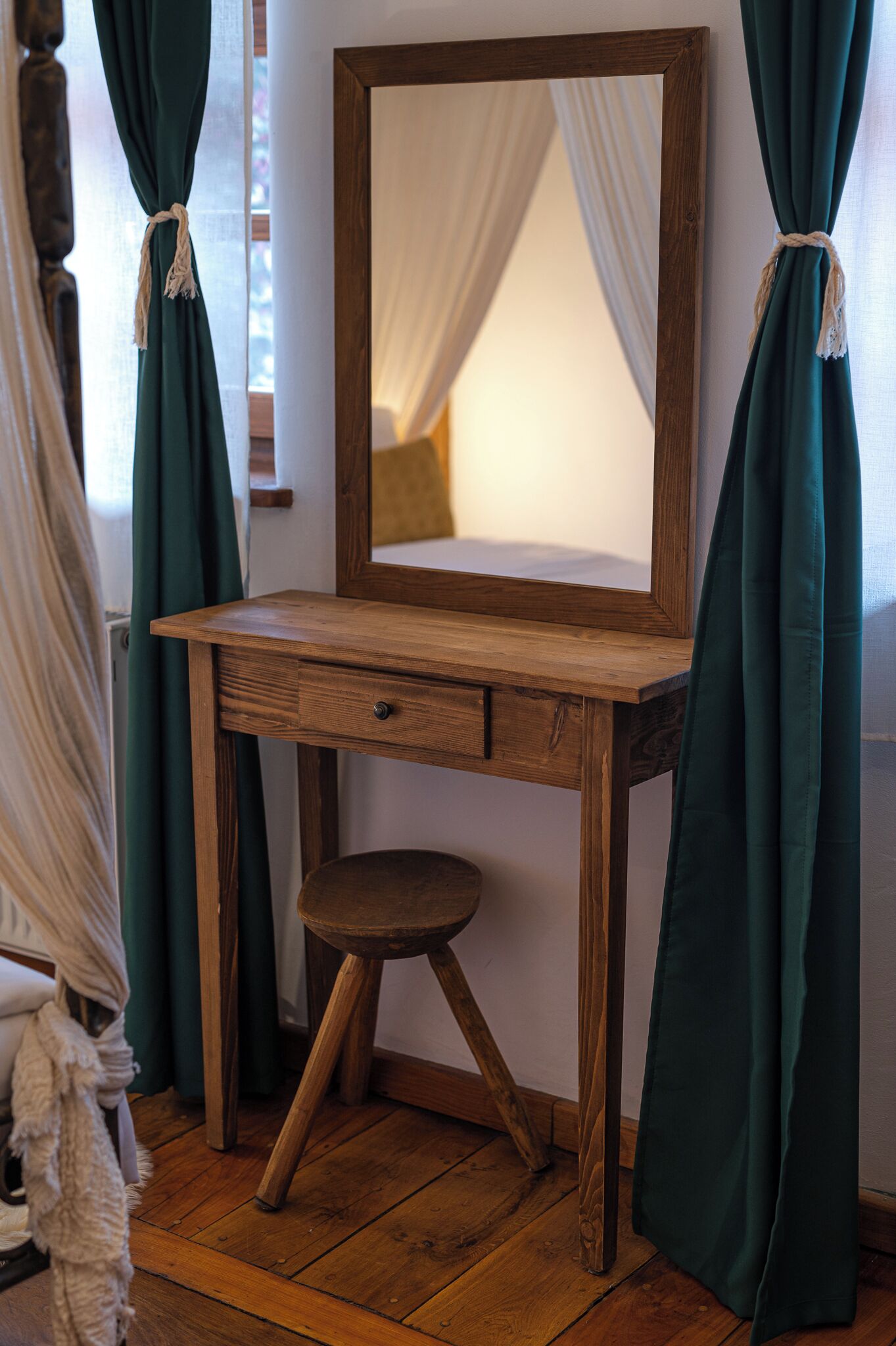 A wooden desk with a mirror, a small stool, and green curtains tied back.