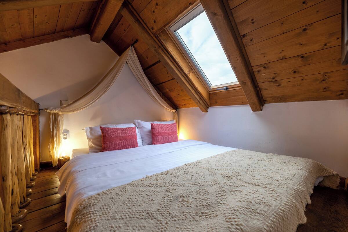 A bedroom with a wooden ceiling, a skylight, a bed with white and red bedding, and a wooden floor.