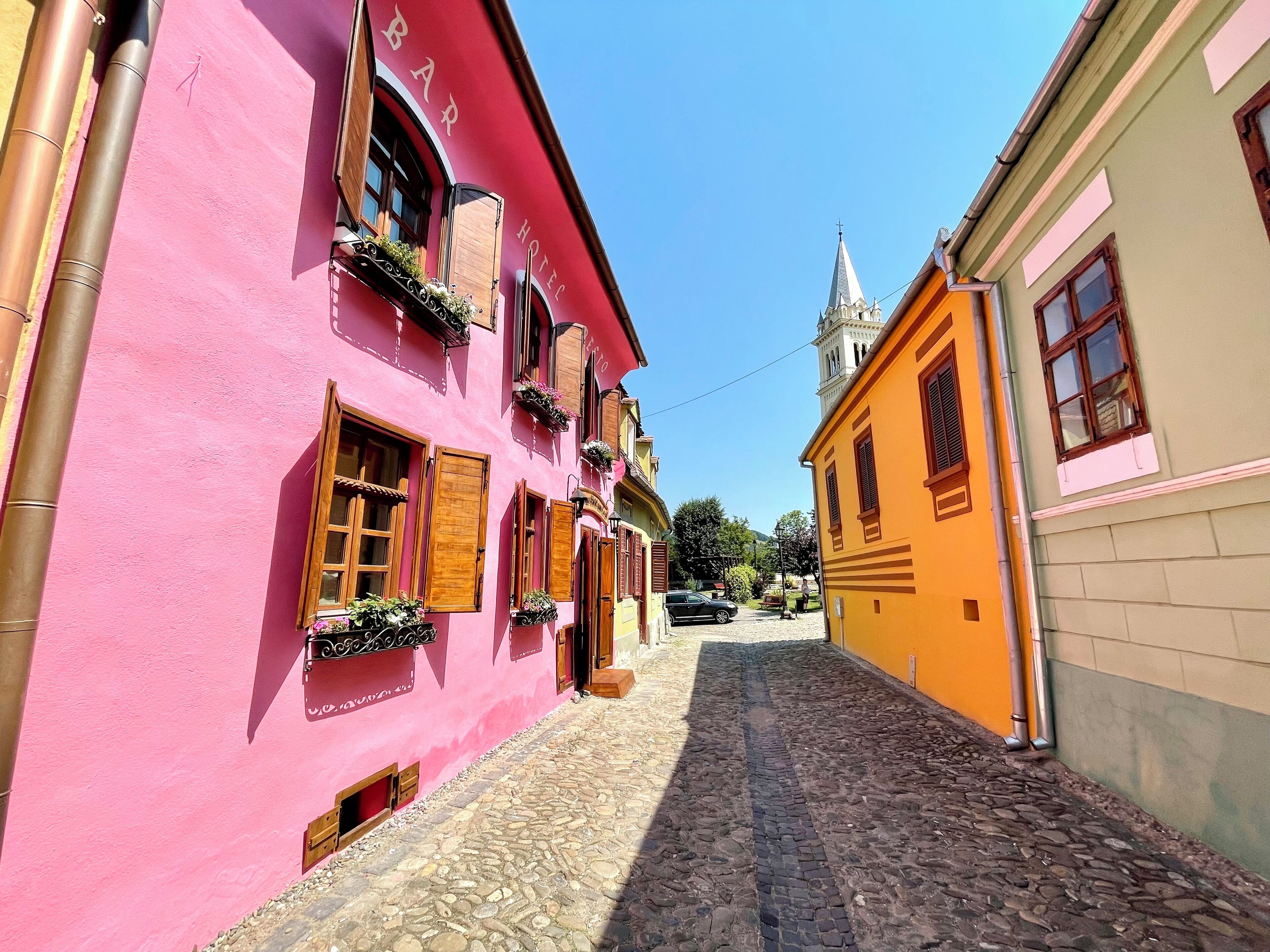 A narrow cobblestone street lined with colorful buildings, including a pink building with wooden shutters and a yellow building with a bell tower.