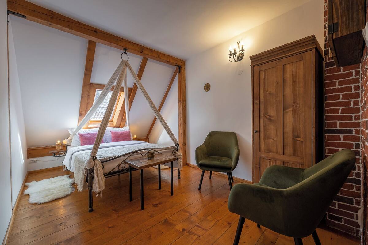 A bedroom with a four-poster bed, a green armchair, a wooden wardrobe, and a brick wall.