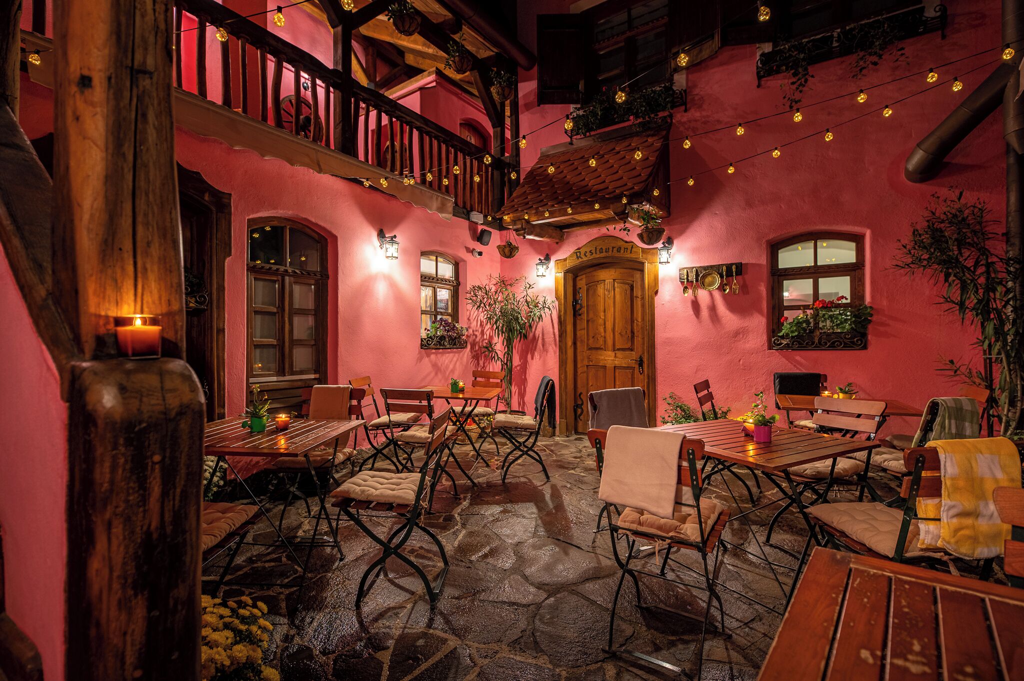 An outdoor dining area with wooden tables and chairs, a stone floor, and a pink building with a wooden door labeled "Restaurant".