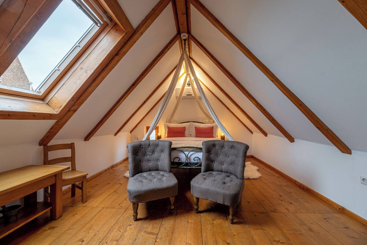 A cozy attic bedroom with a sloped ceiling, a skylight, a bed with red bedding, two grey armchairs, and a wooden bench.