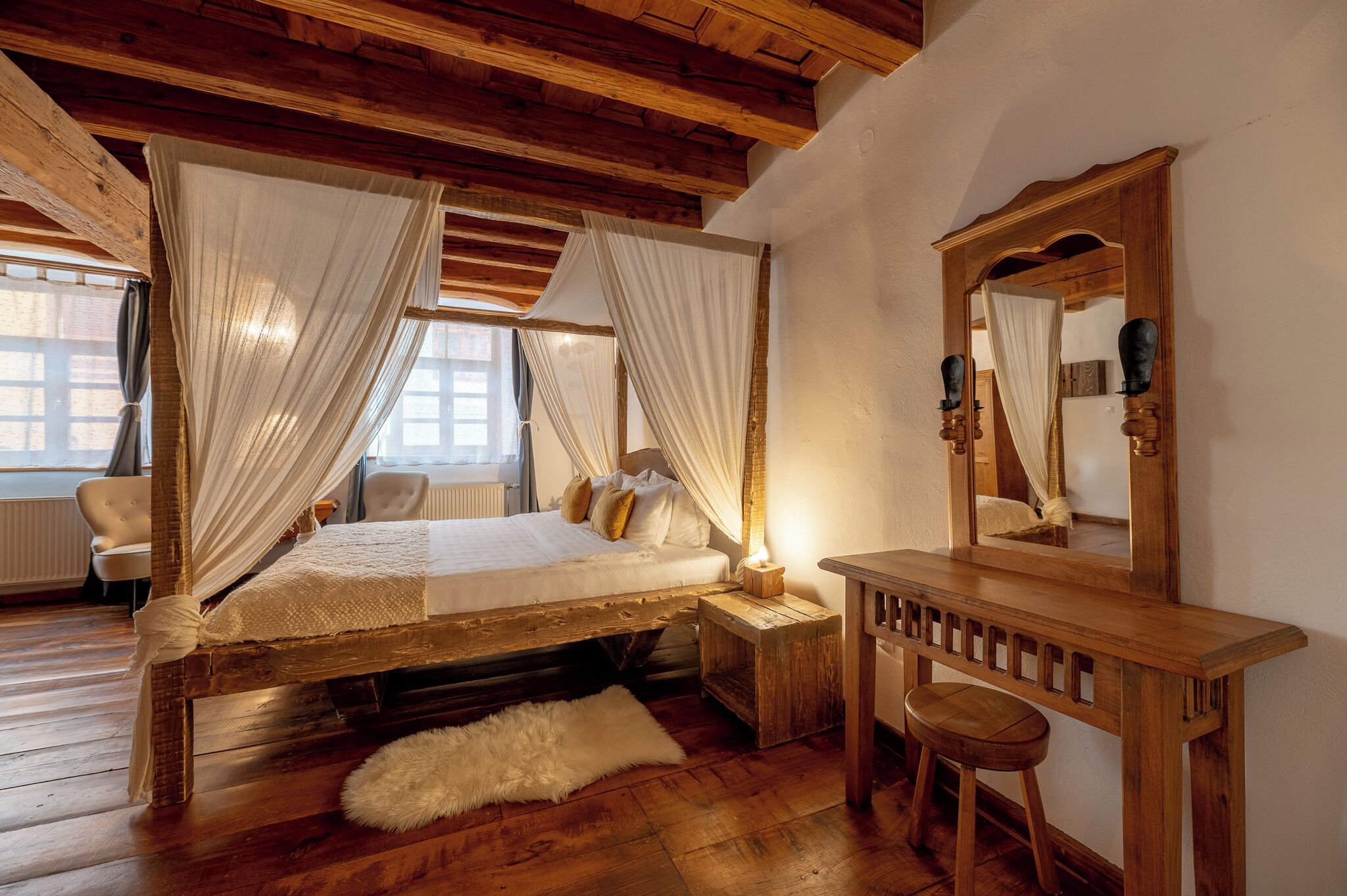 A wooden four-poster bed with white curtains, a wooden nightstand, a mirror, and a white fur rug.
