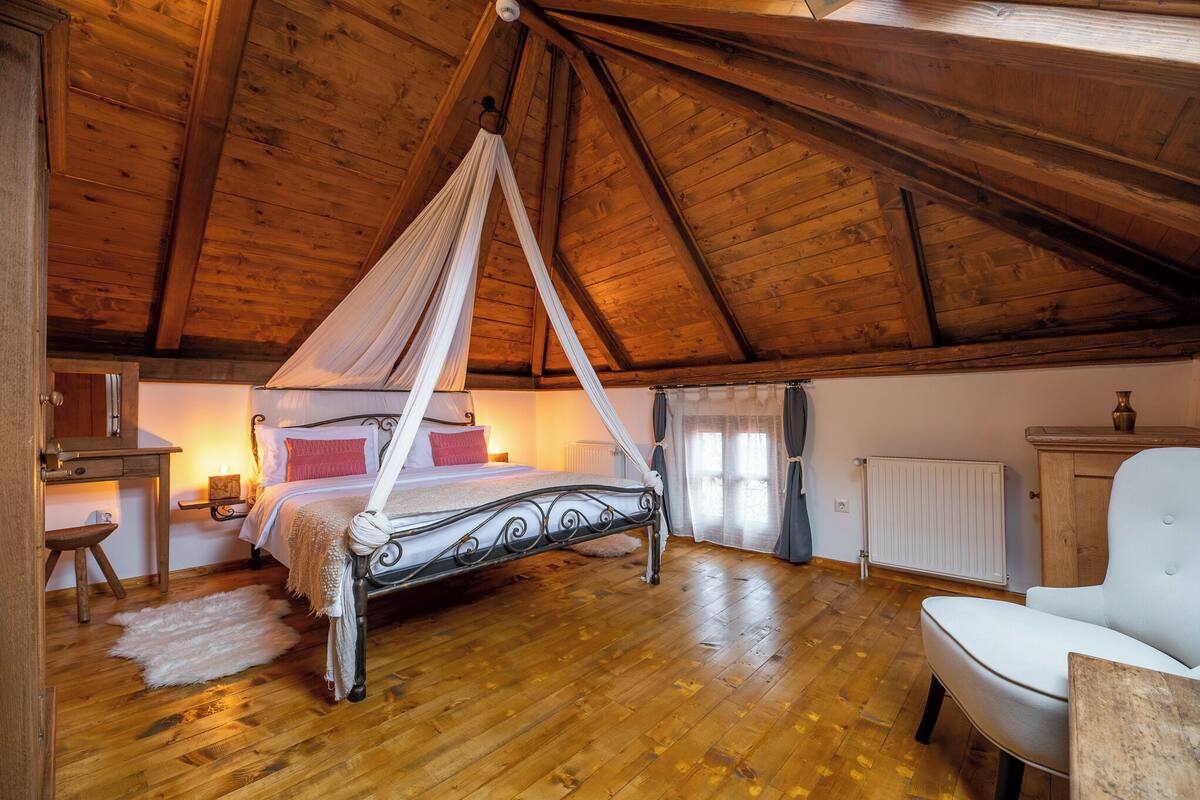 A bedroom with a four-poster bed, wooden flooring, a white chair, and a wooden cabinet.
