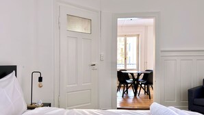 A hotel room with a neatly made bed, a bedside table with a lamp, a dining area with a table and chairs, and a view of the outside through a door. - Cozy & Fully Equipped 2-Bedroom Apartment in Central Zurich (Zürich)