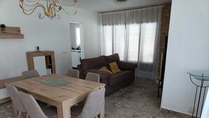 A living room with a wooden dining table, grey chairs, a brown sofa with cushions, a glass coffee table, and a chandelier. - Villa Particular, Confortable (VC)