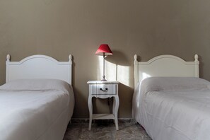A room with two white beds, a small bedside table with a lamp, and a plain wall.