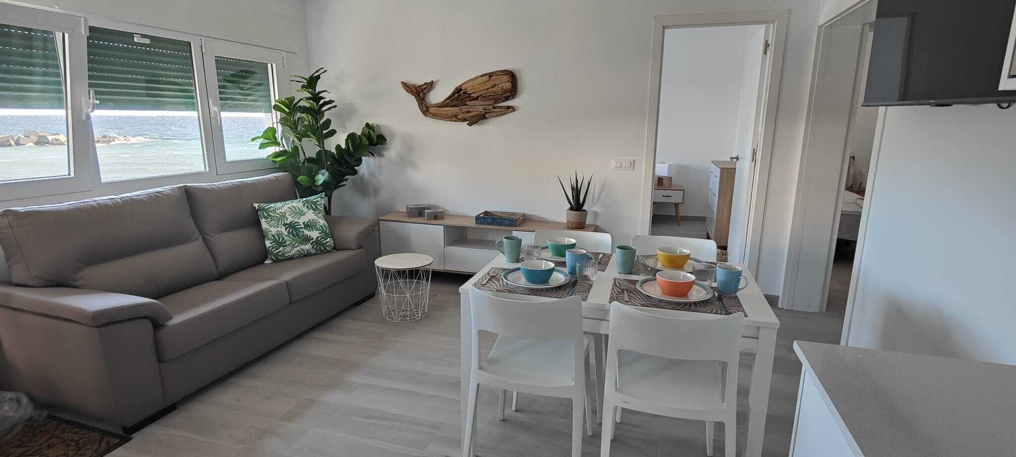 A modern living room with a grey sofa, a dining table set with colorful bowls and cups, a potted plant, and a wooden wall decoration.