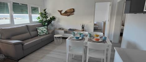 A modern living room with a grey sofa, a dining table set with colorful bowls and cups, a potted plant, and a wooden wall decoration.
