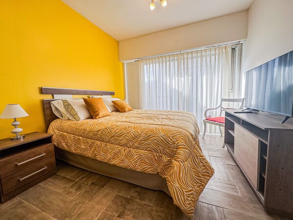 A hotel room with a bed, a desk, a chair, a lamp, and a TV.