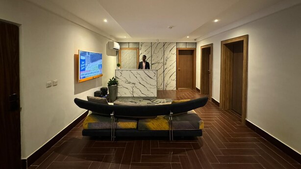 A modern reception area with a marble reception desk, a large flat-screen TV, and a seating area with a sofa and cushions. - Wellaton Suite (Abuja)
