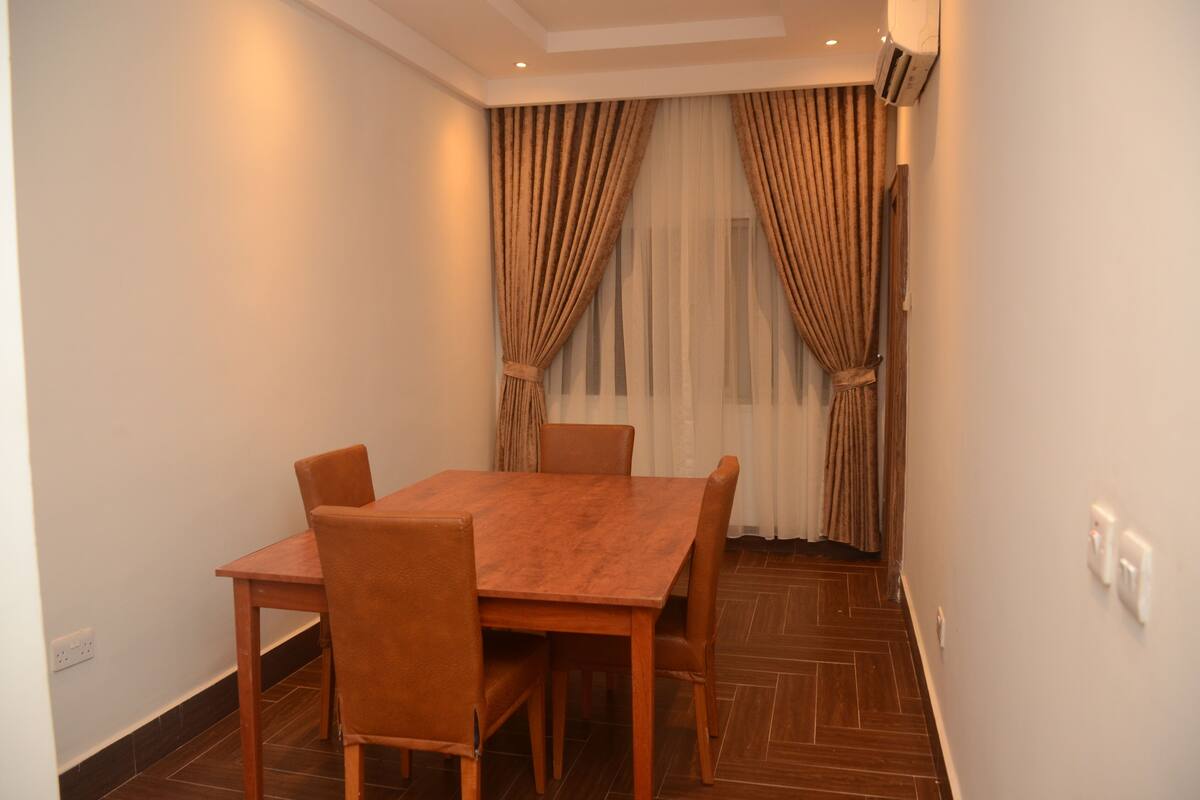 A room with a wooden table and four chairs, a window with curtains, and a tiled floor.
