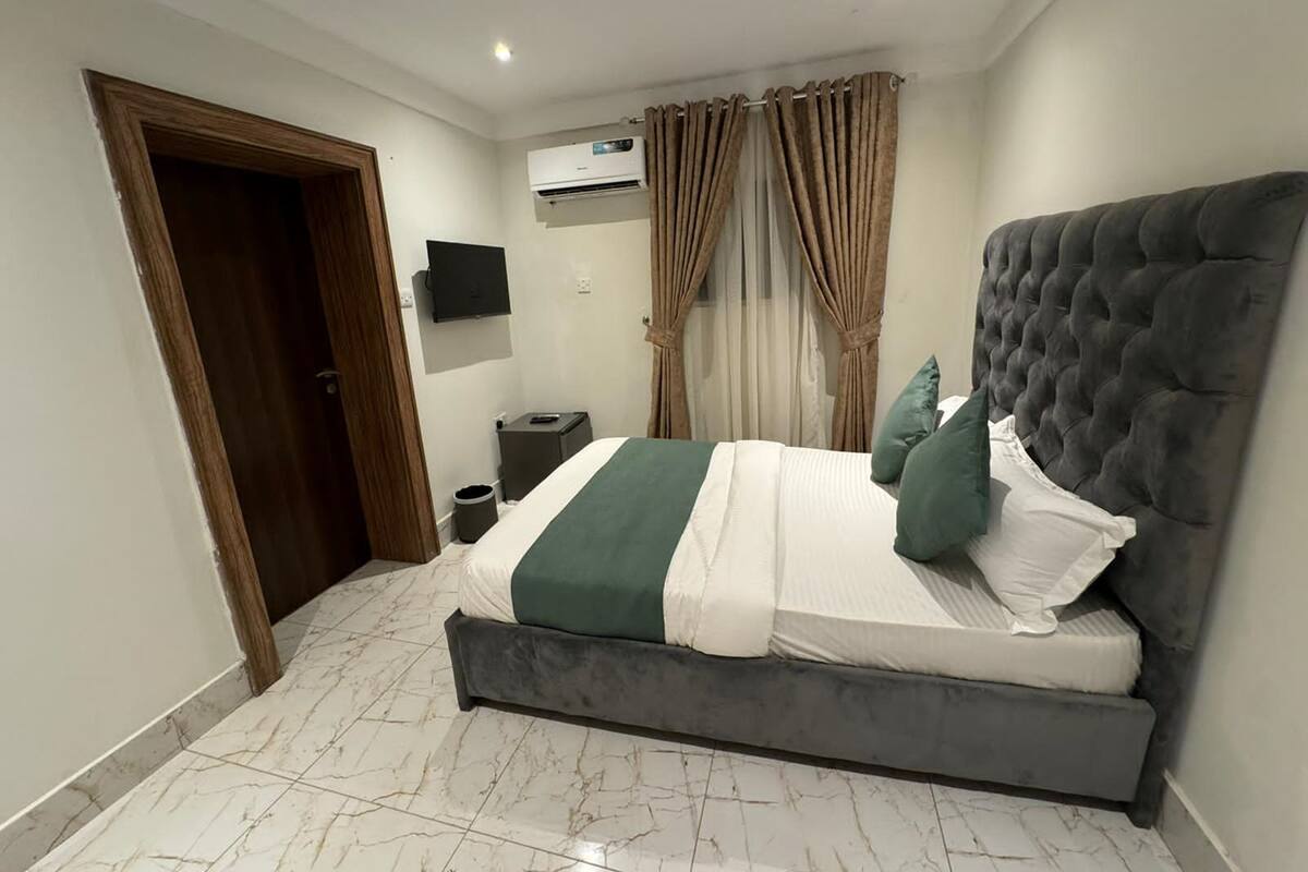 A bedroom with a large bed, a TV mounted on the wall, an air conditioning unit, and a door leading to another room.