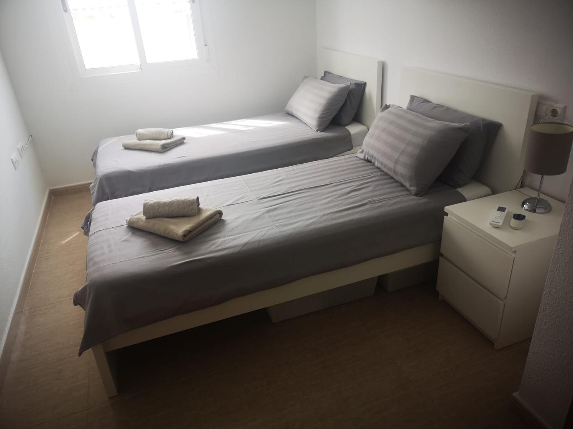 A twin bedroom with two beds, a nightstand with a lamp, and a window providing natural light.