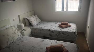 Une chambre à coucher avec deux lits, chacun agrémenté d’une couvre-lit et de coussins à motifs, une table de chevet avec une lampe et une fenêtre donnant sur des immeubles.