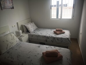 Ein Schlafzimmer mit zwei Betten, jeweils mit gemusterten Bettdecken und Kissen, einem Nachttisch mit einer Lampe und einem Fenster mit Blick auf Gebäude.