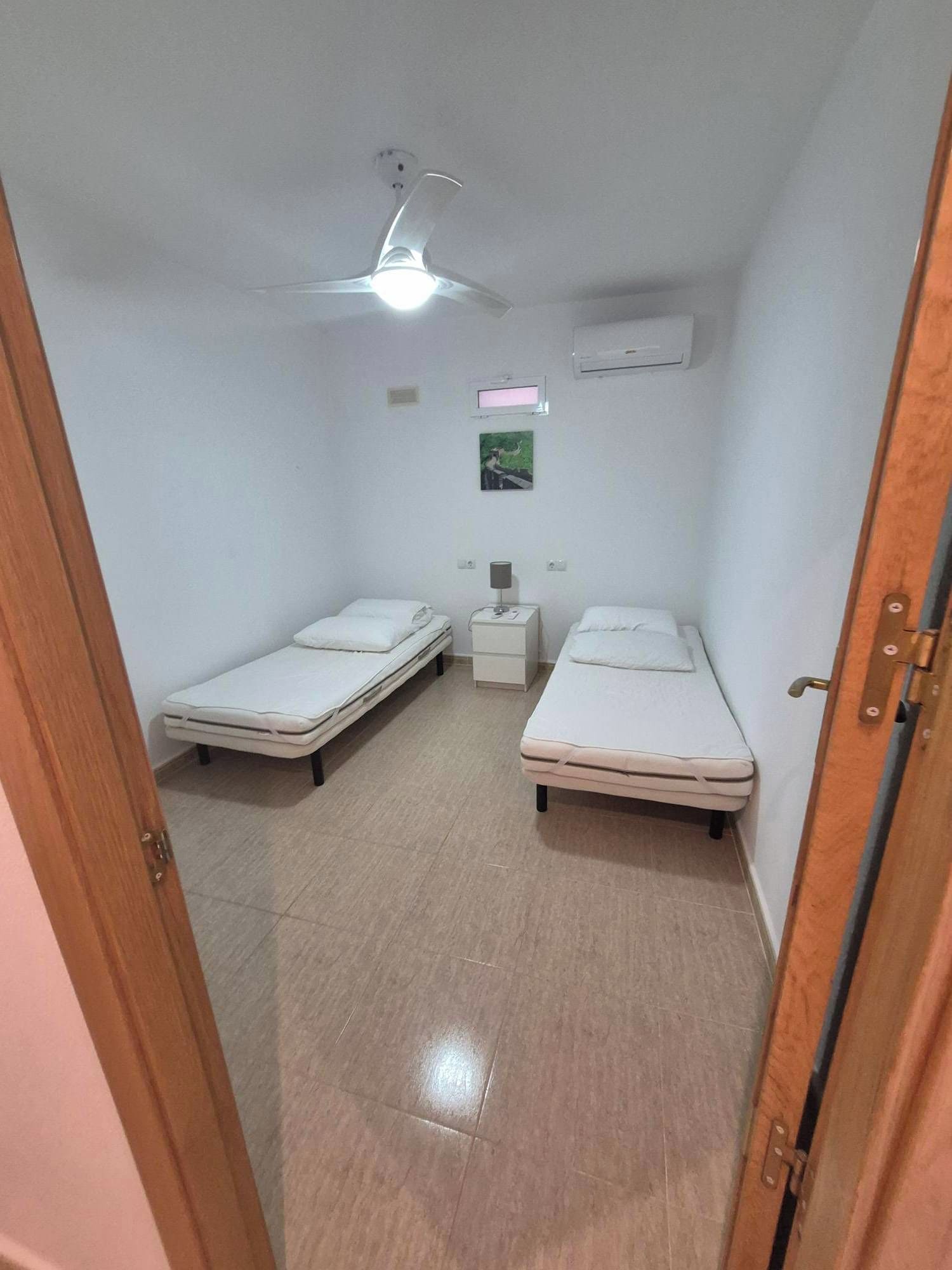 A small, narrow room with two single beds, a nightstand, a wall-mounted air conditioner, and a ceiling fan.