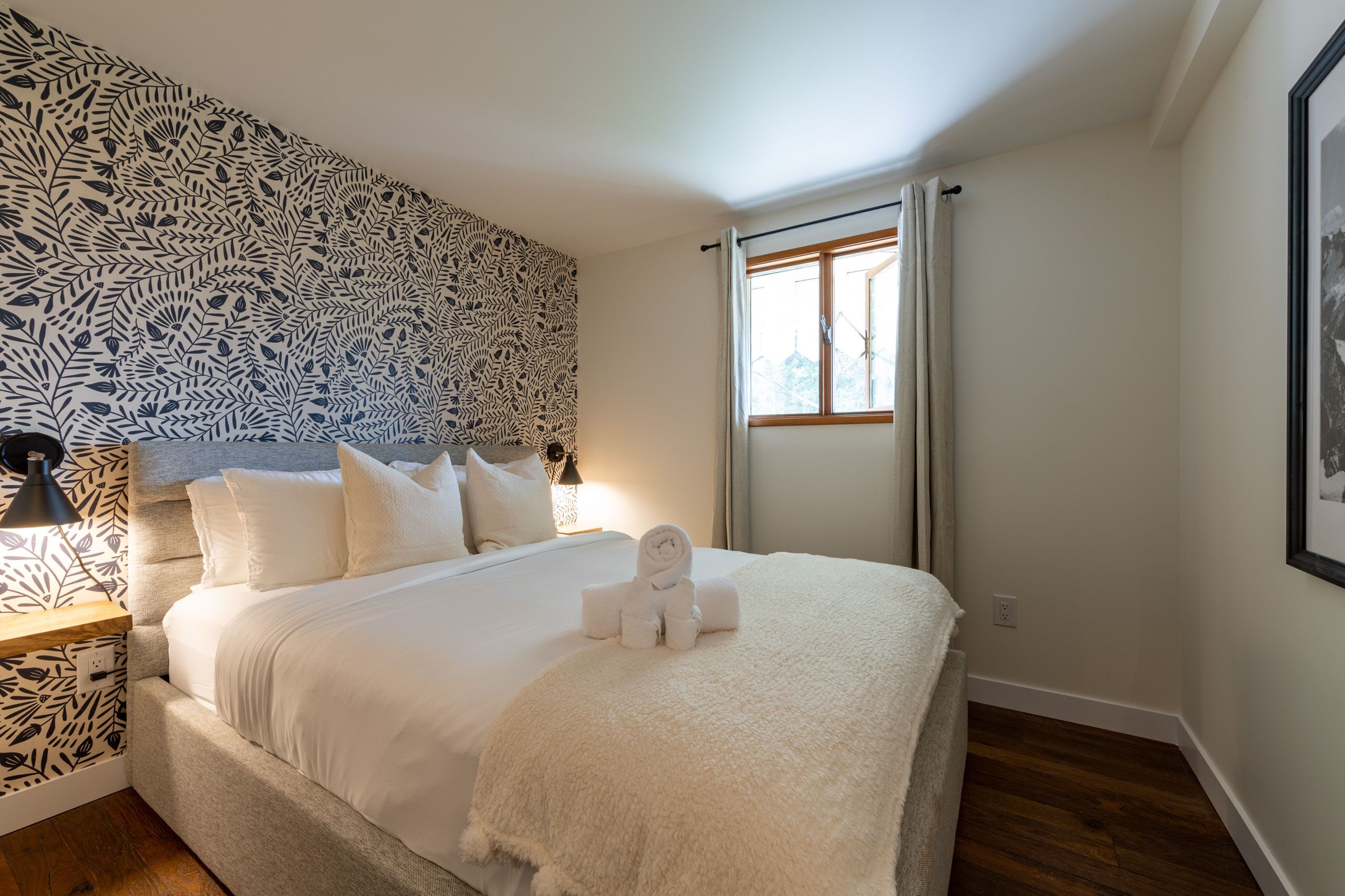 A neatly made bed with white linens and a rolled towel, a bedside lamp, and a patterned wall.