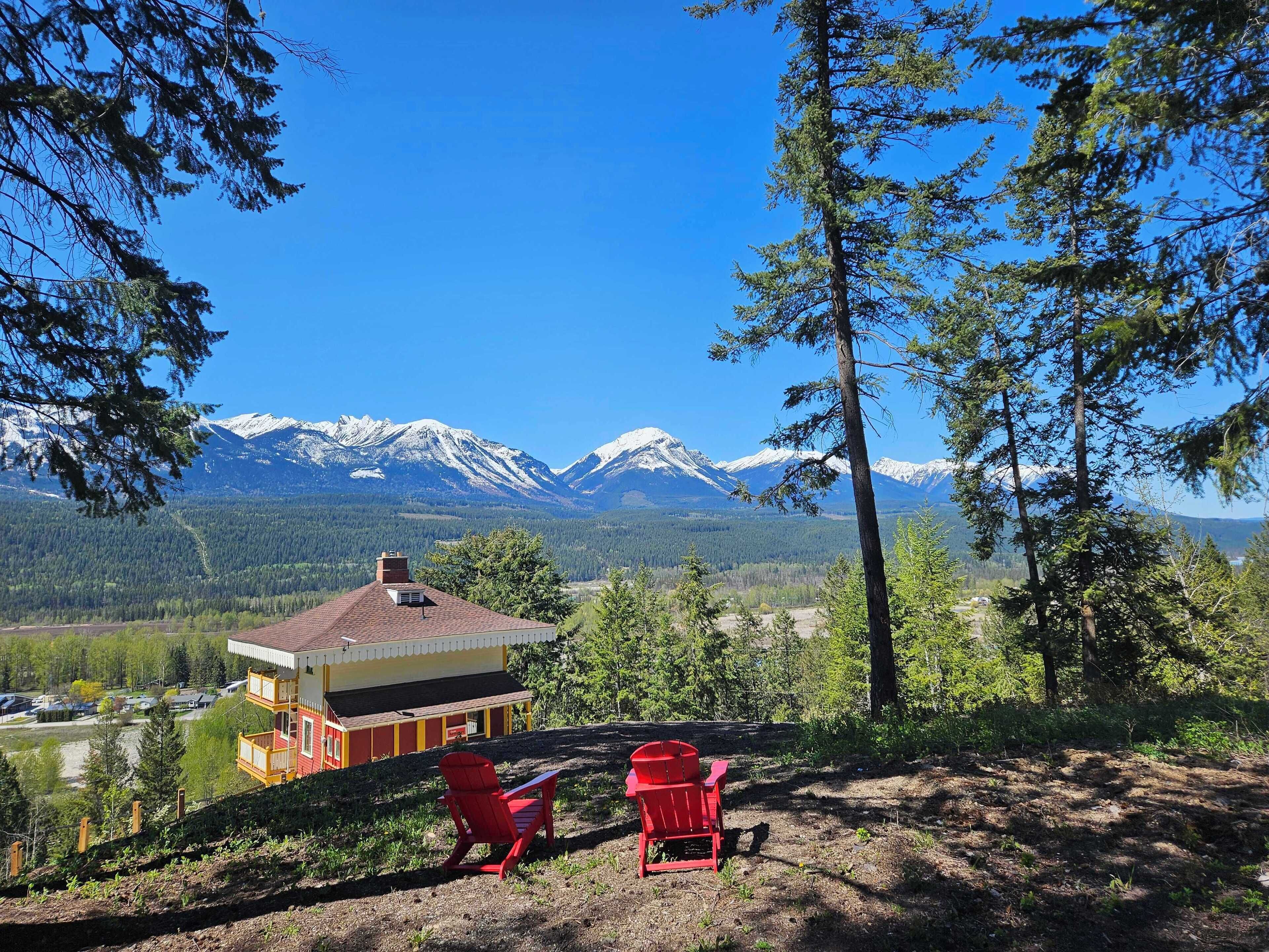 A cabin with a red roof and yellow walls, two red Adirondack chairs, and a view of snow-capped mountains.