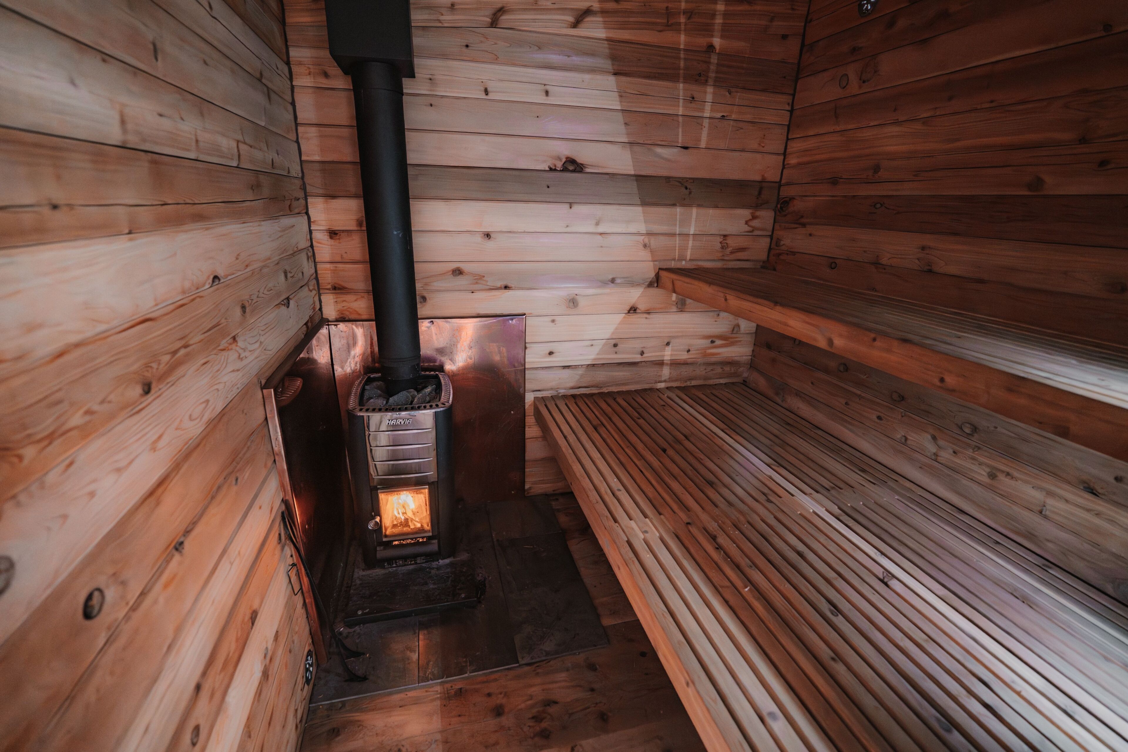 A wooden cabin interior with a wood-burning stove, benches, and a fireplace.