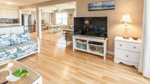 A living room with a sofa, TV, and a lamp. There's a dining area with chairs and a table. The kitchen is visible with cabinets and appliances. - In Duck with Community Pool & Beach Access (Duck)