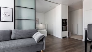 A modern living room with a grey sofa, a kitchen with white cabinets, and a large window. - Stylish studio near the National Library (Minsk)