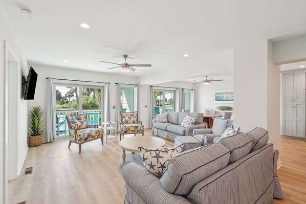A spacious living room with a large sofa, a coffee table, and a TV mounted on the wall. There are two ceiling fans, a dining table with chairs, and a kitchen area visible through an open door. - Oceanfront Escape: Brand-New Sea Cabin with Stunning Upgrades (Isle of Palms)