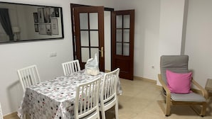 A dining area with a table set for four, white chairs, a wooden door with glass panels, and a chair with a pink cushion.
