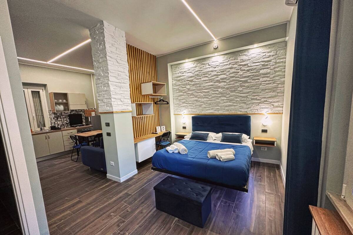 A modern hotel room with a large bed, a desk, and a kitchenette.