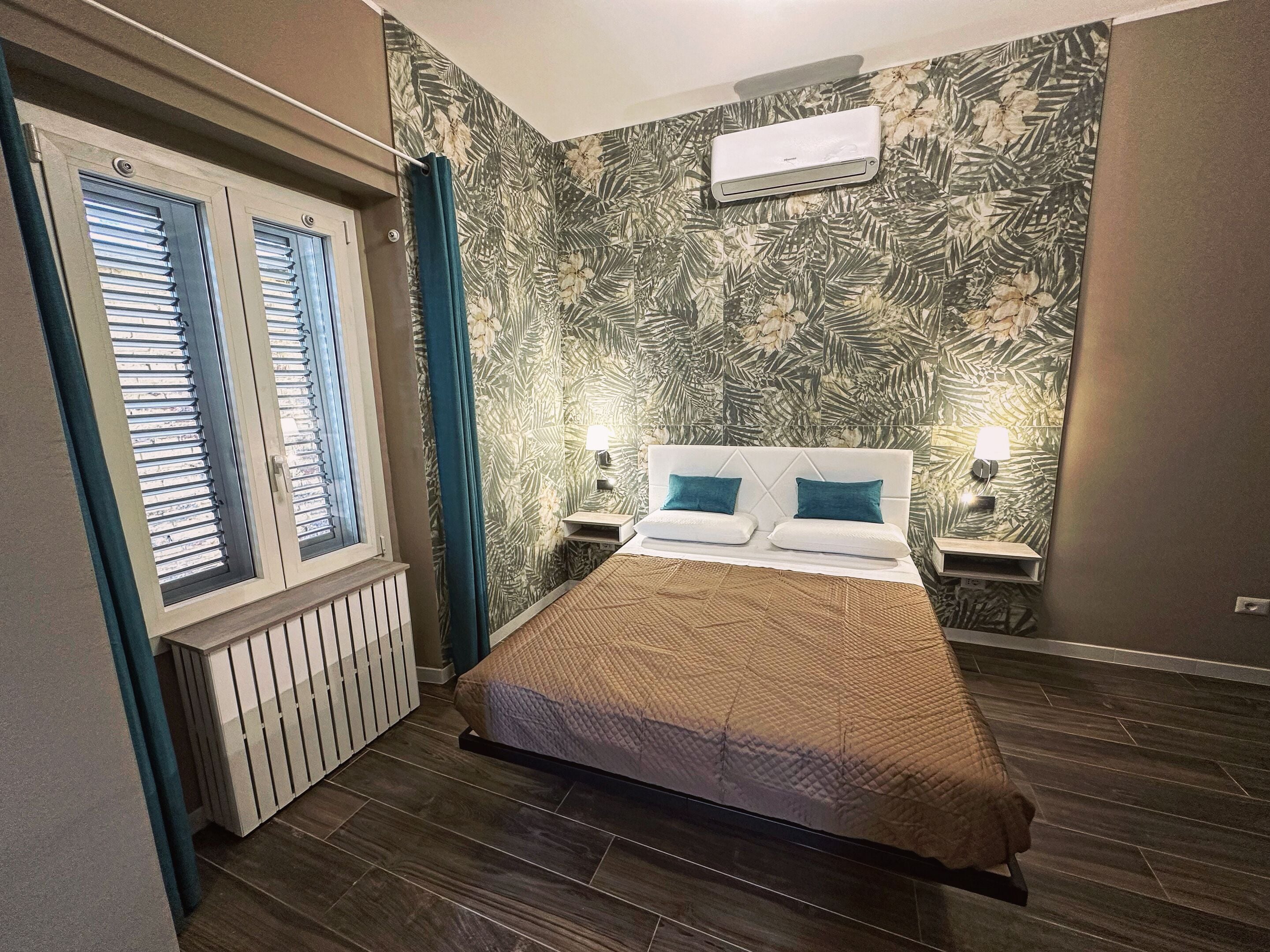 A bedroom with a bed, two bedside lamps, a window with blinds, a radiator, and a wall-mounted air conditioner.