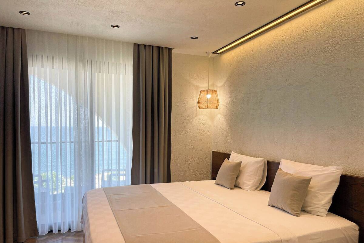 A hotel room with a large bed, a view of the sea through sheer curtains, a wall-mounted light fixture, and a wooden headboard.