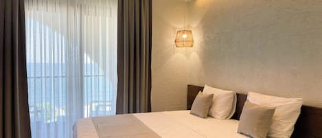 A hotel room with a large bed, a view of the sea through sheer curtains, a wall-mounted light fixture, and a wooden headboard.