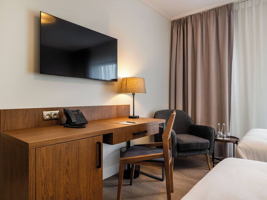 A hotel room with a wooden desk, a chair, a bed, a TV mounted on the wall, and a lamp.