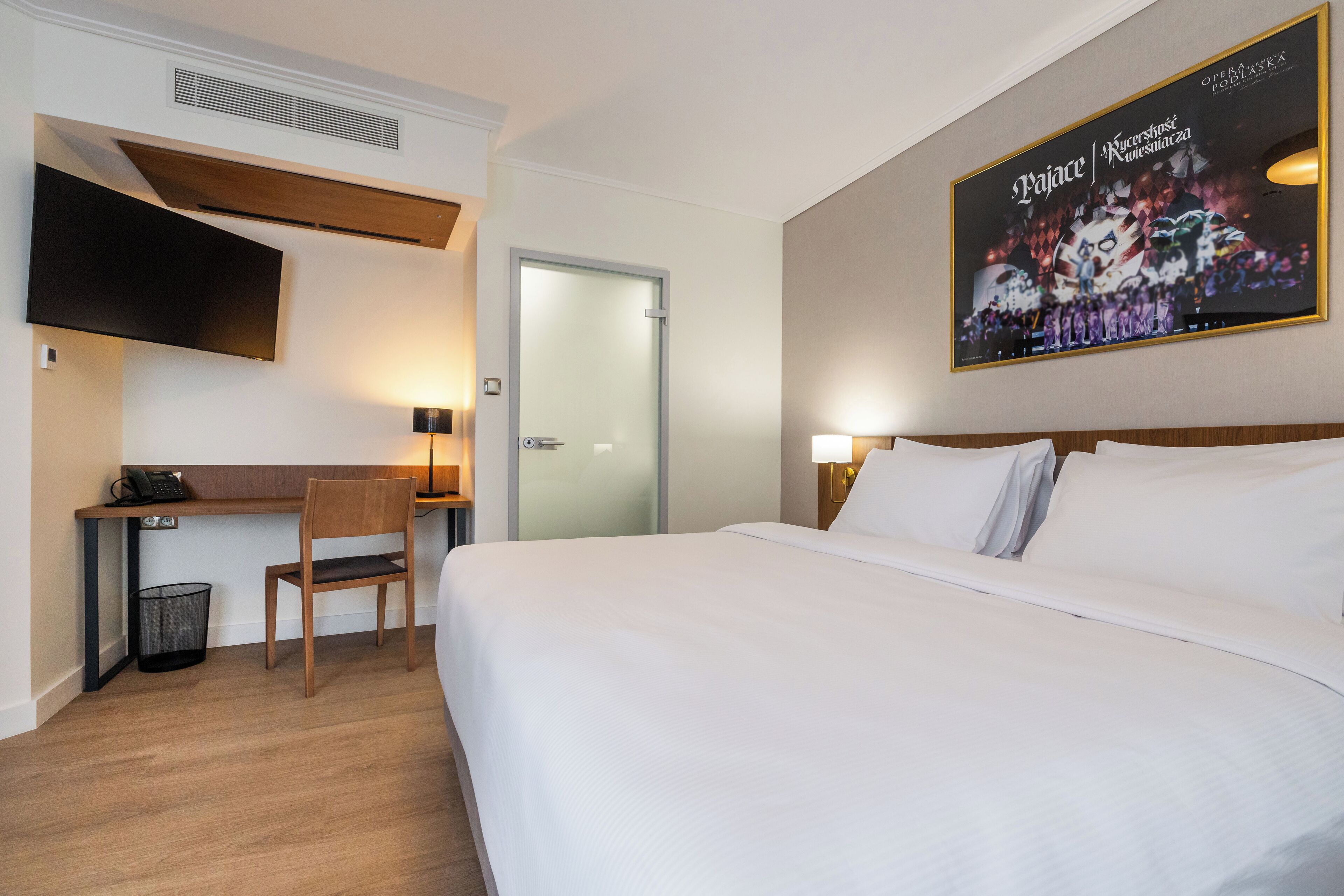 Quarto de hotel com uma cama, uma escrivaninha, uma cadeira, uma TV fixada na parede e um cartaz.