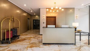 Zona d'entrada d'hotel amb sòl de marbre, taulell de recepció, una làmpada penjant i un carro de roba amb malates.