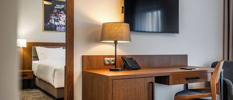 A hotel room with a wooden desk, a telephone, a lamp, a bed, and a TV mounted on the wall.