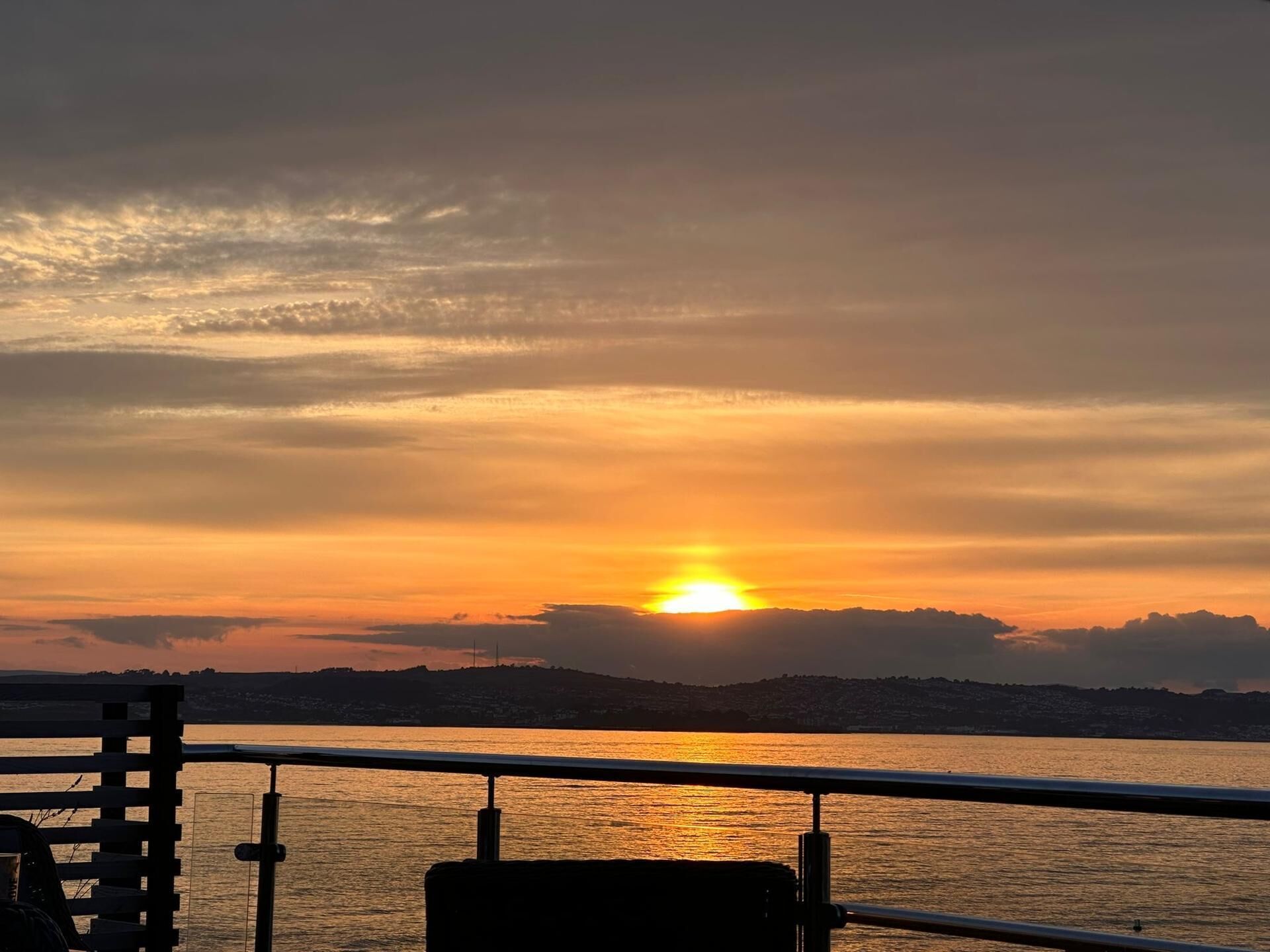 Un atardecer sobre una masa de agua con una costa distante y una barandilla en primer plano.