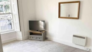 A room with a grey sofa, a TV on a wooden stand, a white radiator, and a window with a view of the street. - Torquay, 1 Bedroom, Sleeps 4, Free Parking, Torbay (Torquay)
