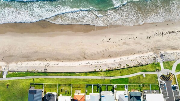 Vista aérea de una zona costera con playa, un sendero cubierto de hierba y edificios con diversas estructuras.