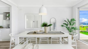 Un comedor moderno con decoración blanca, una mesa grande, sillas, una lámpara colgante y vistas al exterior.