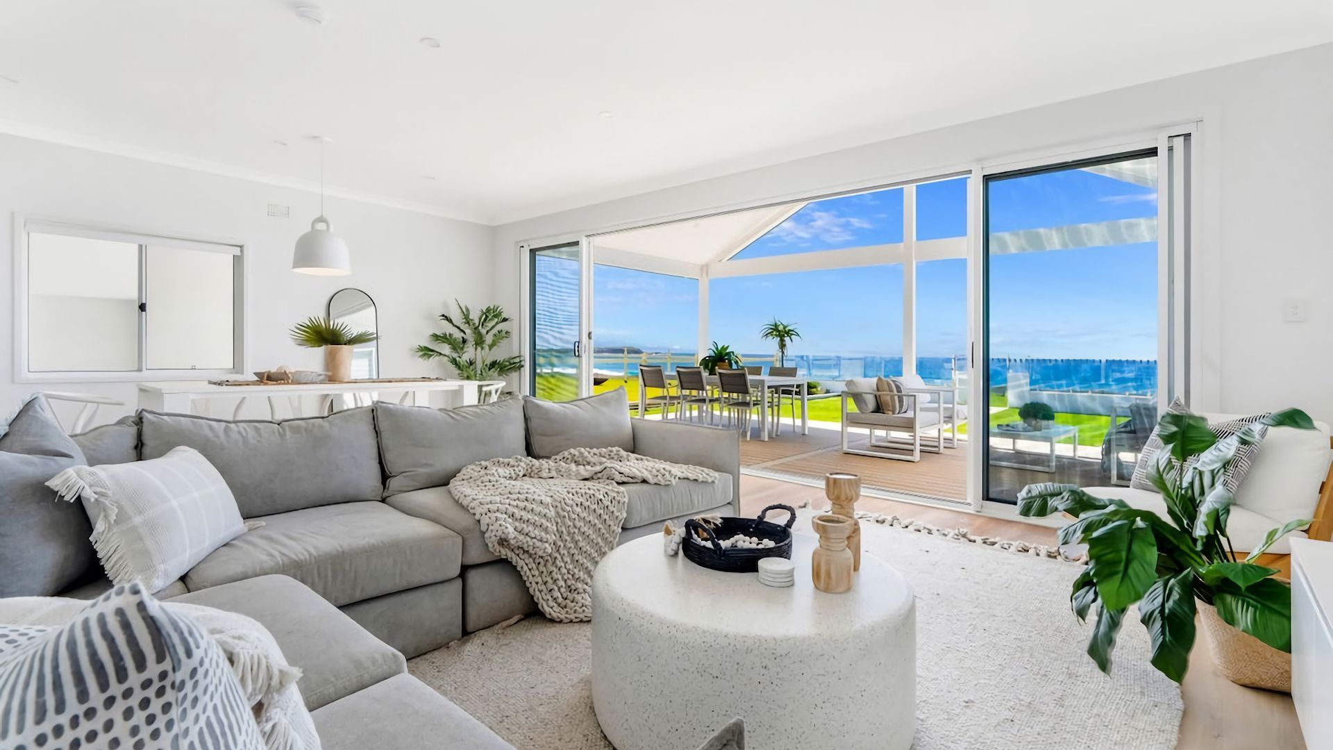 A modern living room with a large grey sofa, a round coffee table, and a view of the ocean through large glass doors.