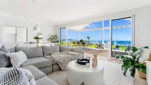 A modern living room with a large grey sofa, a round coffee table, and a view of the ocean through large glass doors. - La Palma Beach House ~ Charming coastal retreat (Warilla)