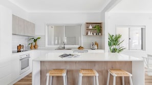 A modern kitchen with white cabinetry, a large island with stools, a built-in oven, and a window with a view. - La Palma Beach House ~ Charming coastal retreat (Warilla)