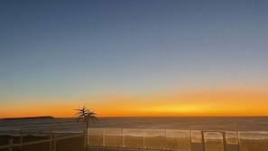 Vista del atardecer desde un balcón con vista al océano, con una palmera y una baranda de madera.