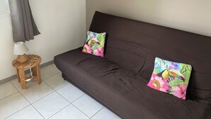 Um sofá marrom escuro com duas almofadas coloridas, uma com design de tucano e outra floral, um banquinho de madeira com uma cesta trançada e um abajur, e uma janela com cortinas cinzas.
