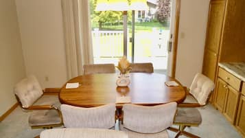 A dining room with a round table, six chairs, a vase with dried flowers, and a view of a backyard through a glass door.